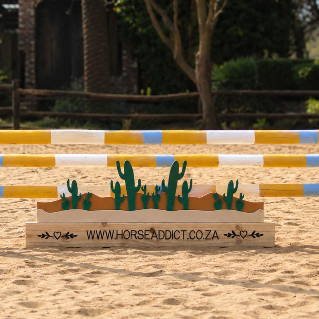 Cactus wooden show jump filler positioned beneath yellow white and blue jump poles in a sand arena