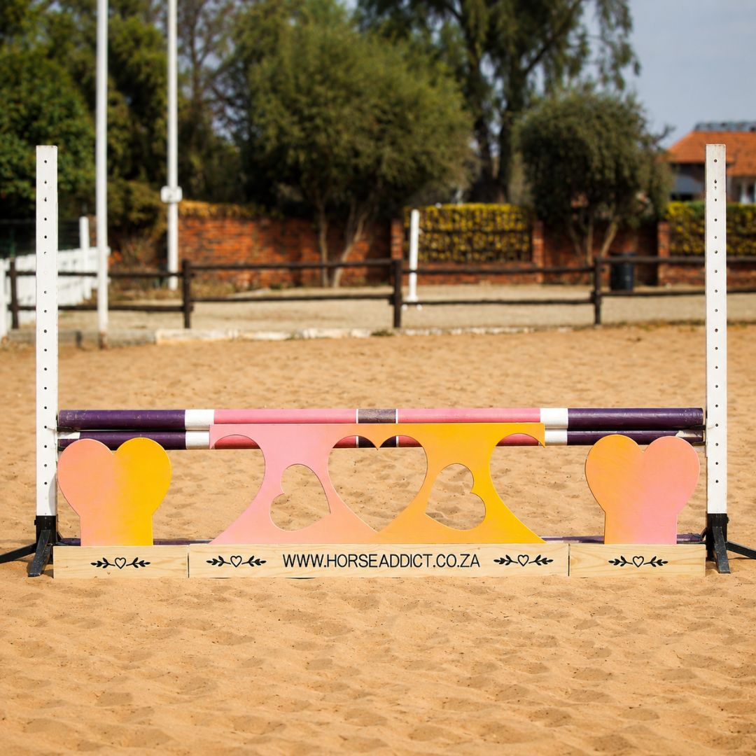 Colourful sunset heart wooden jump filler set positioned beneath purple and pink show jumping poles in a sand arena